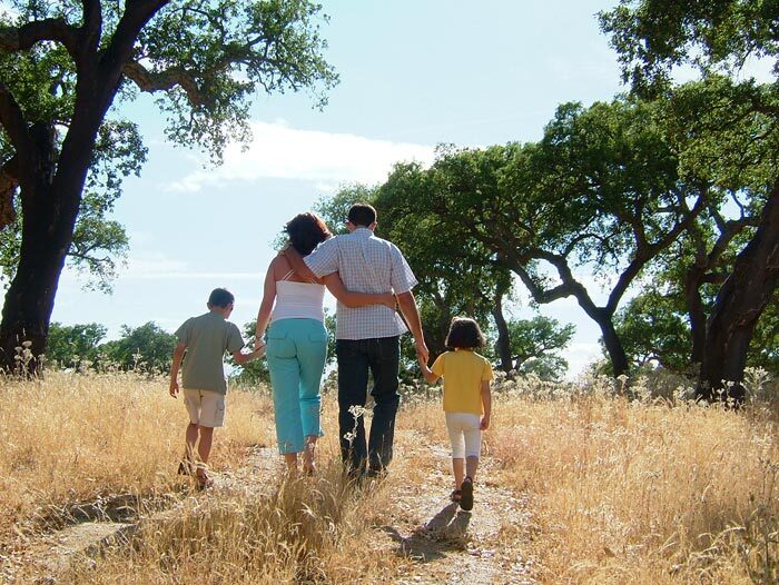 Fam&iacute;lia a caminhar de m&atilde;os dadas por um trilho entre sobreiros, numa paisagem de vegeta&ccedil;&atilde;o seca.